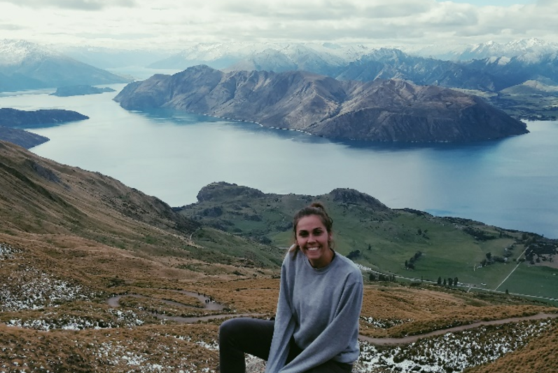 Female student sitting on a mountain (Roys Peak in New Zealand) with water and an island in the background.