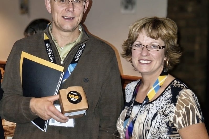 A man holding a notebook and a small box and a woman holding a glass are smiling at the camera.