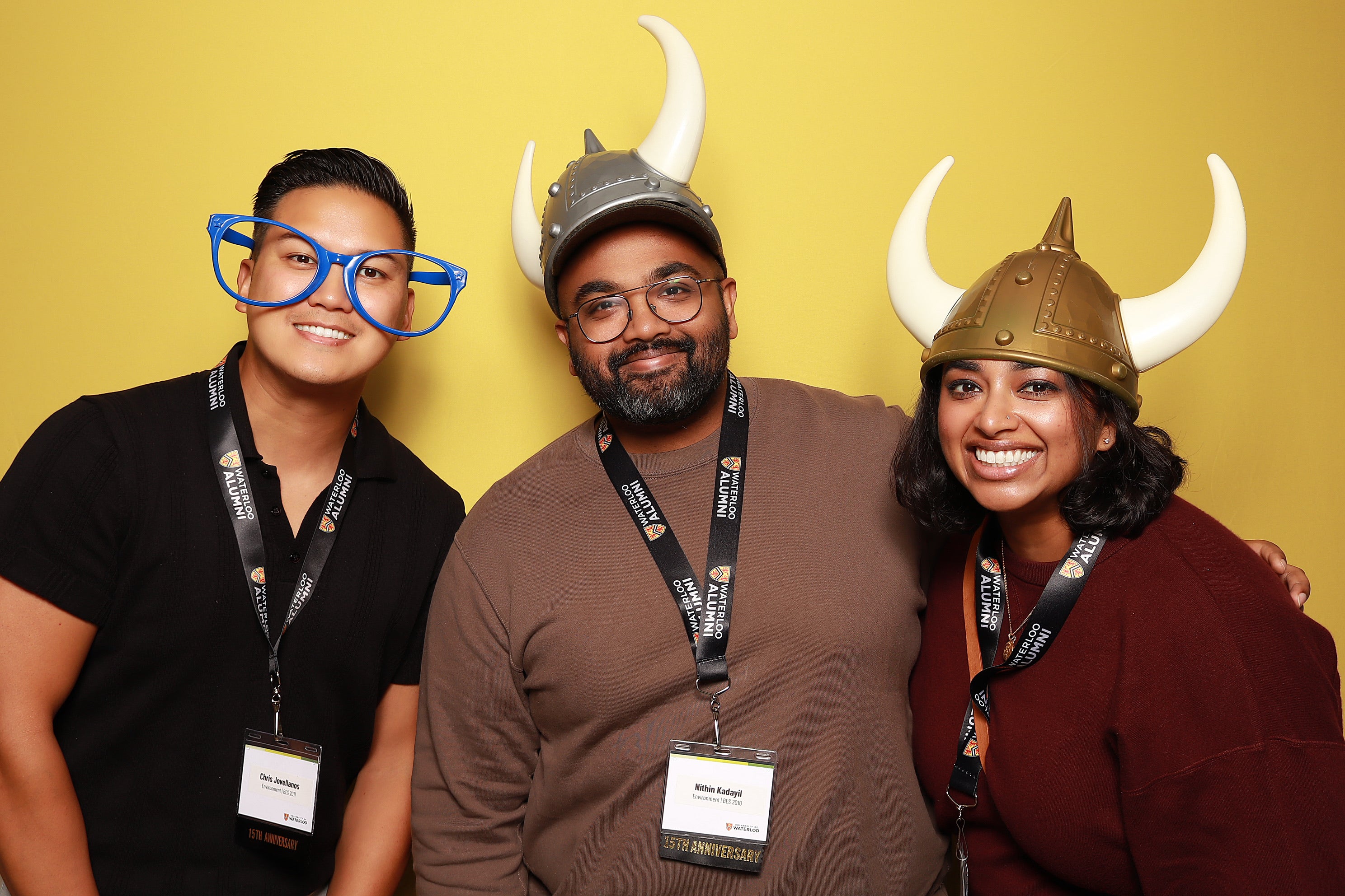 Three people with oversized glasses and viking hats smile in front of a yellow background.