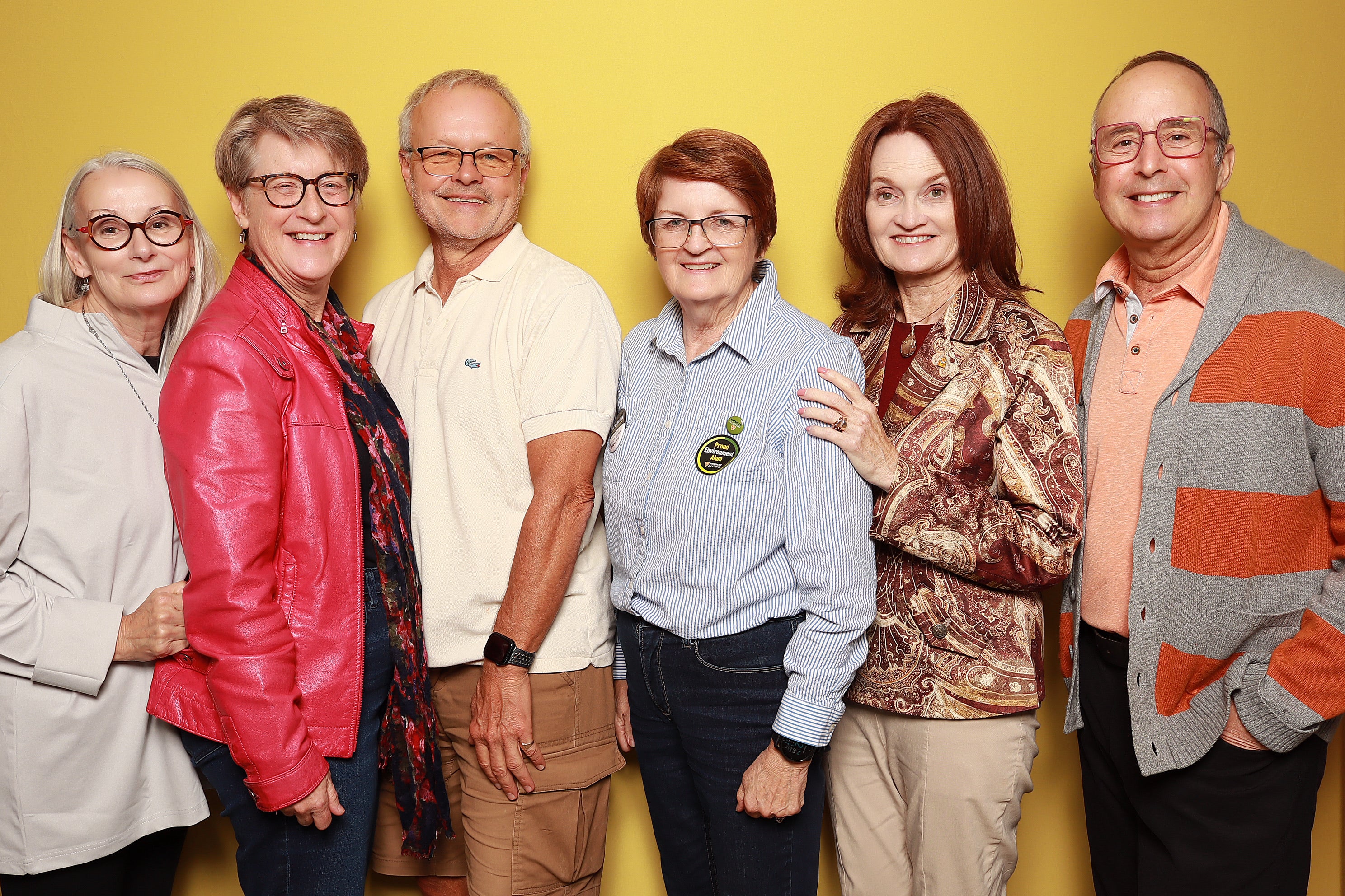 A group of six adults standing side by side against a yellow background, dressed in various styles and colors.