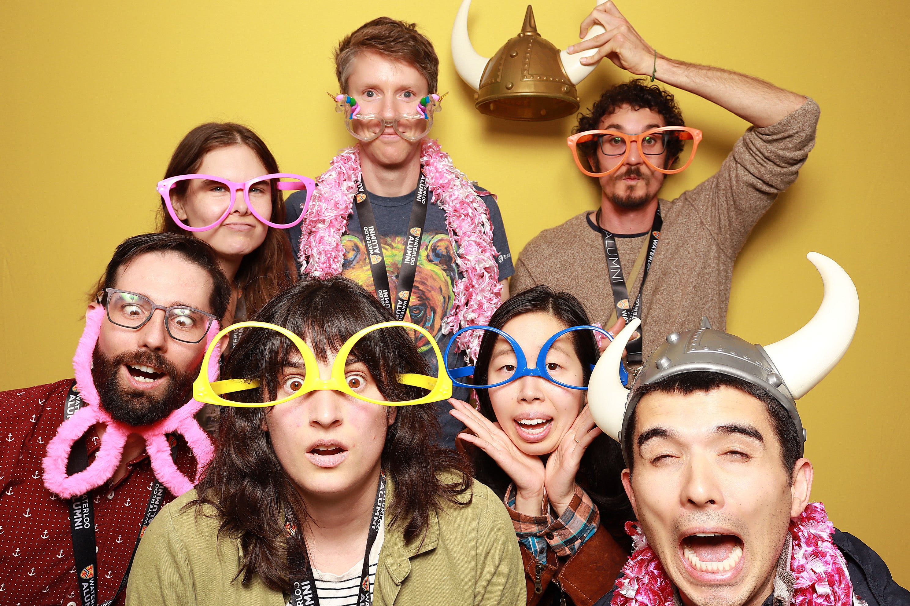 A group of seven people wearing silly glasses, colorful accessories, and playful expressions, posing against a yellow background.