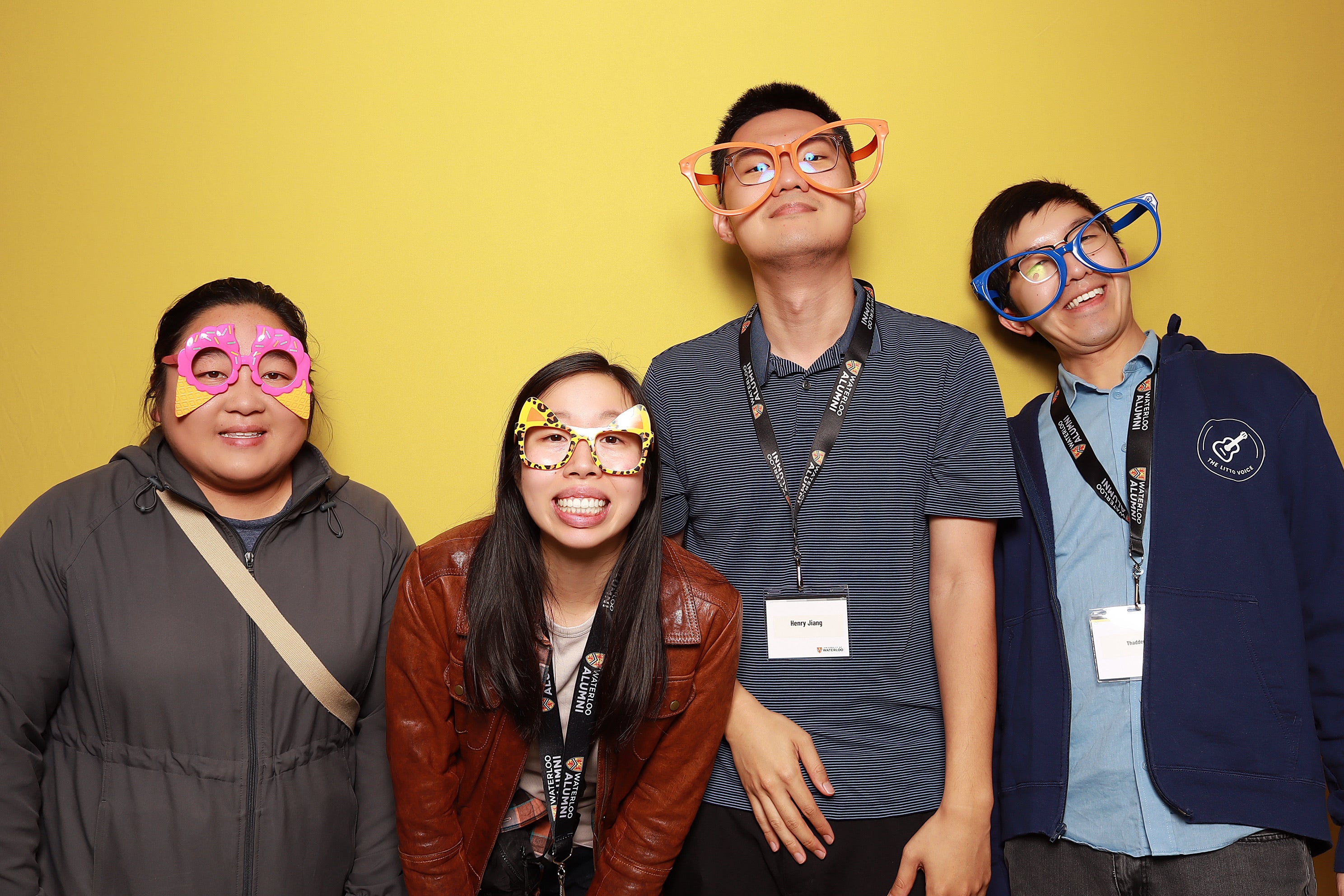 Four people with silly glasses smile in front of a yellow background.
