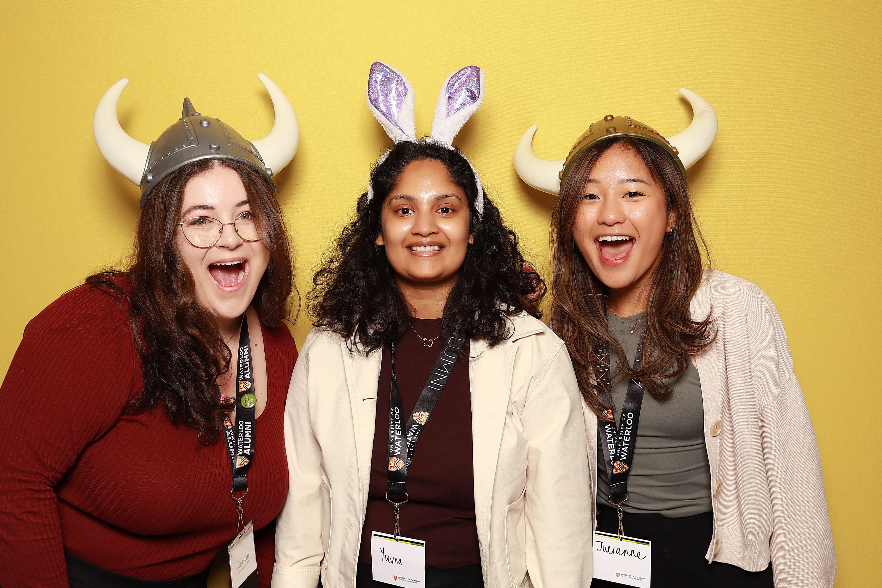 Three people wearing fun hats and accessories stand in front of a yellow backdrop, smiling.