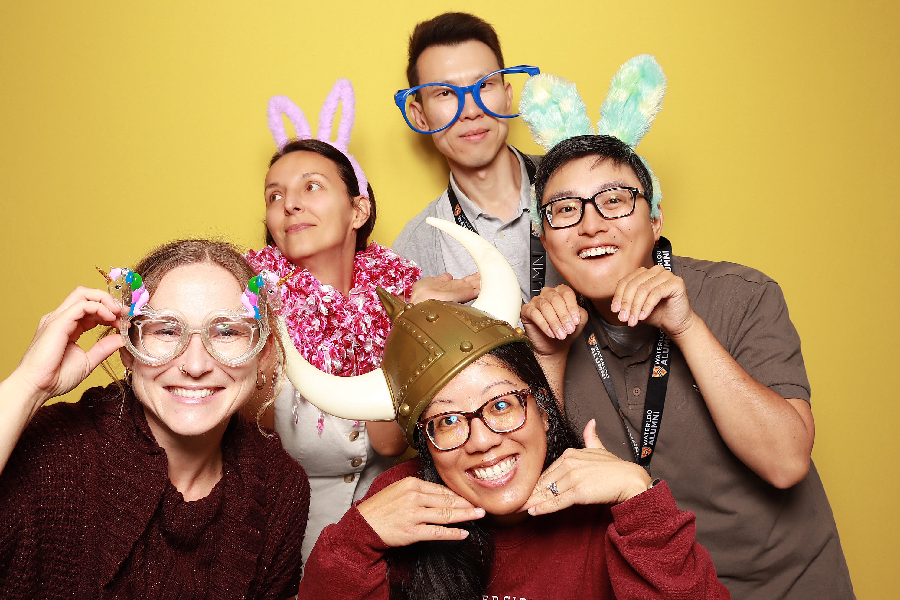 A group of five people pose with silly props in front of a yellow wall. 