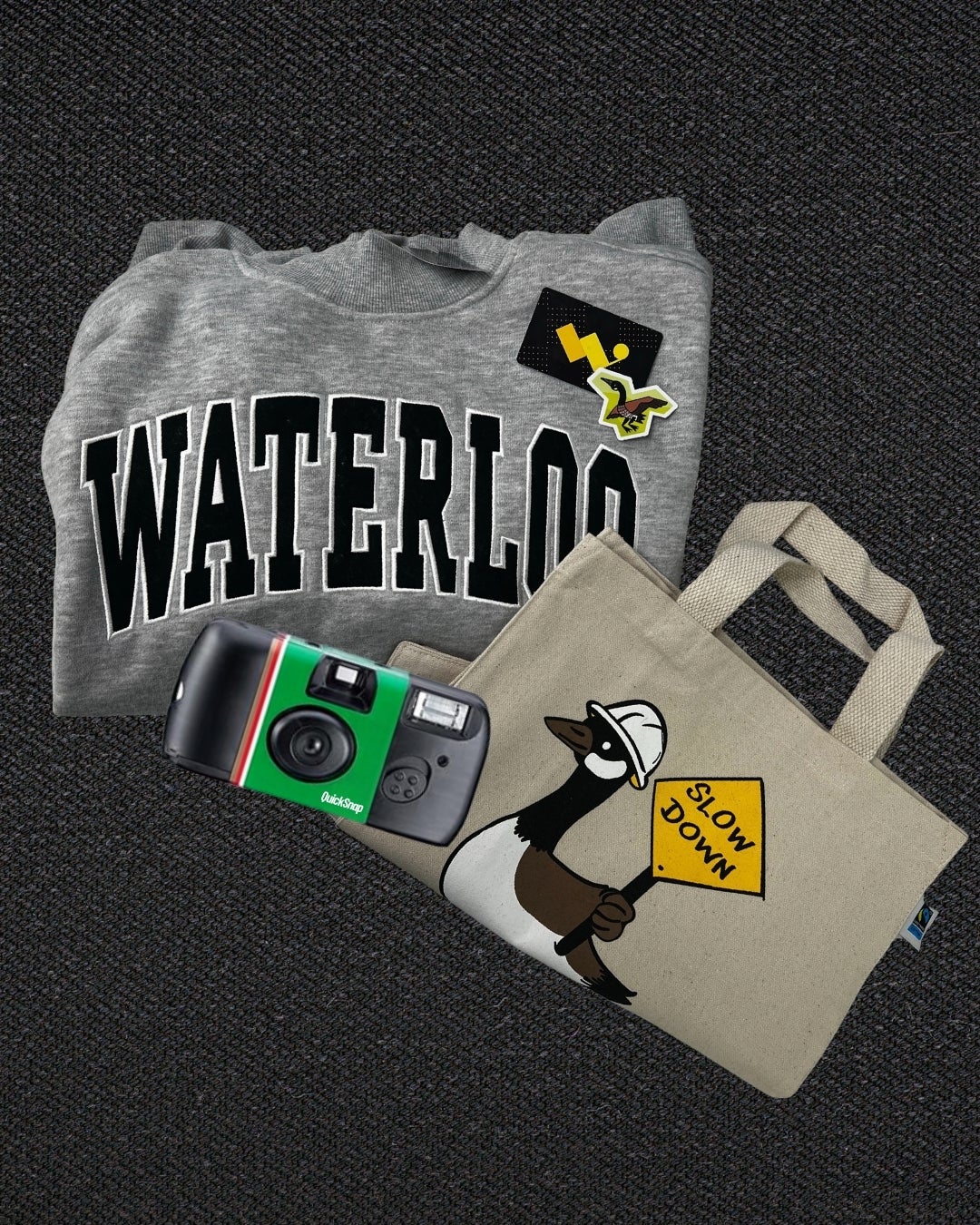 A sweater embroidered with Waterloo, a tote bag, gift card, disposable camera and canadian goose sticker placed on a black carpet