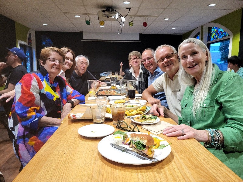 A group of people sitting at a long table in a restaurant, smiling and enjoying their meal.