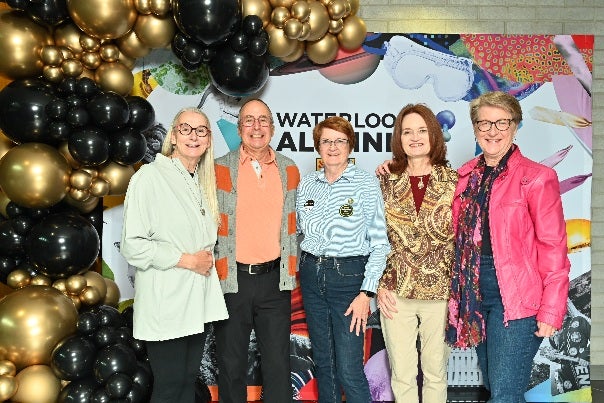 A group of five people posing together in front of a colorful backdrop that says "WATERLOO ALUMNI," decorated with balloons.