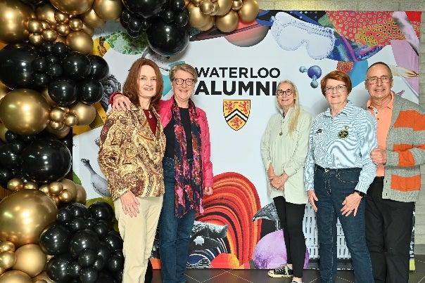 A group of five people posing together in front of a colorful backdrop that says "WATERLOO ALUMNI," decorated with balloons.
