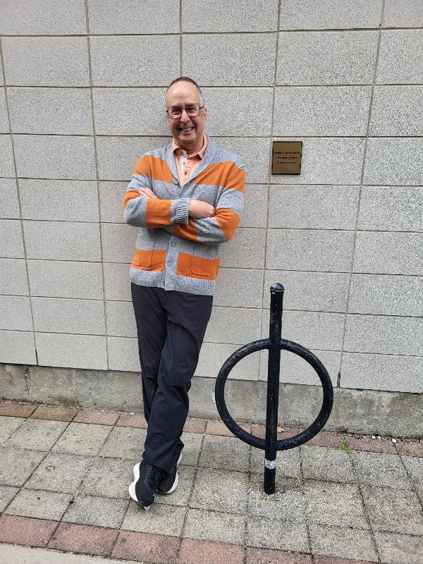 A man with glasses and a beard stands casually next to a circular signpost, wearing an orange and gray striped sweater, black pants, and sneakers, against a gray wall.