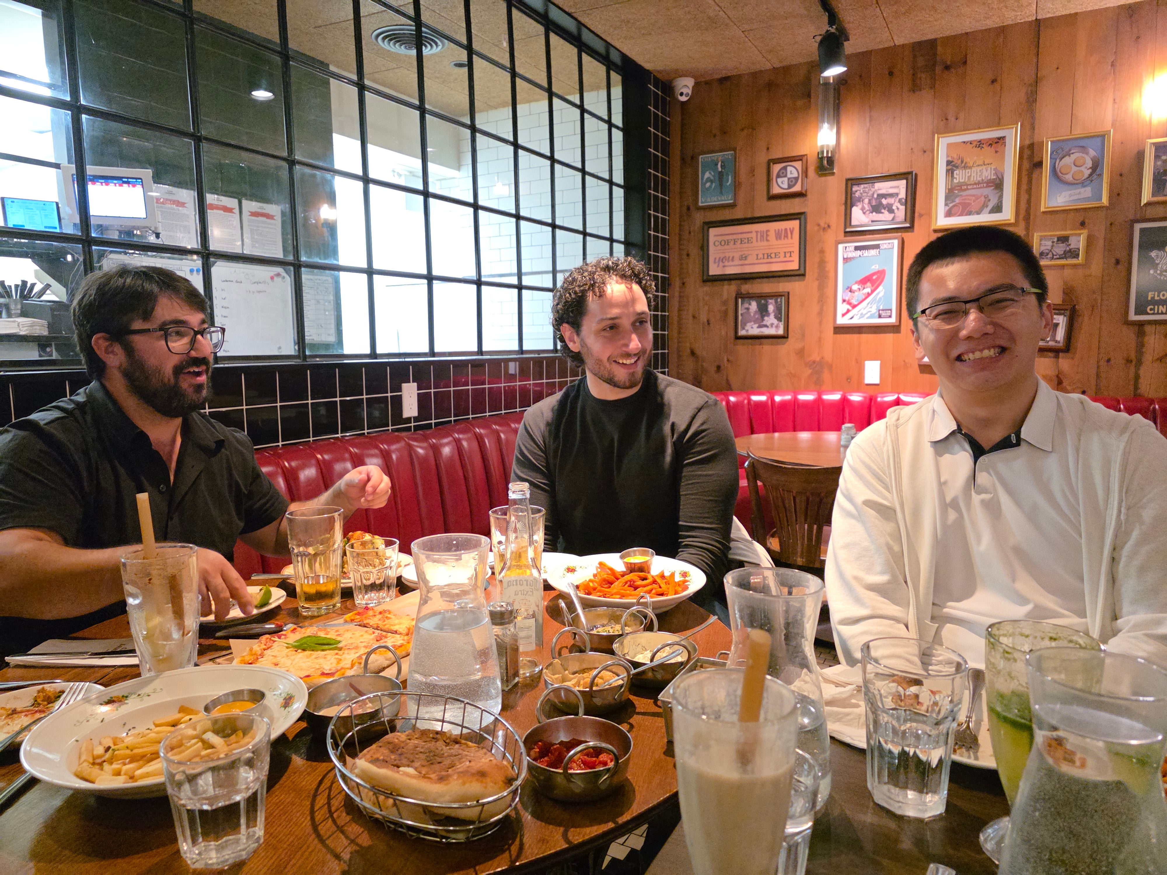 Three people are sitting at a restaurant table filled with food, smiling. 