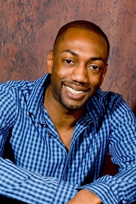 Caustan DeRiggs: young black man leaning casually forward with hands crossed, wearing a blue checkered shirt
