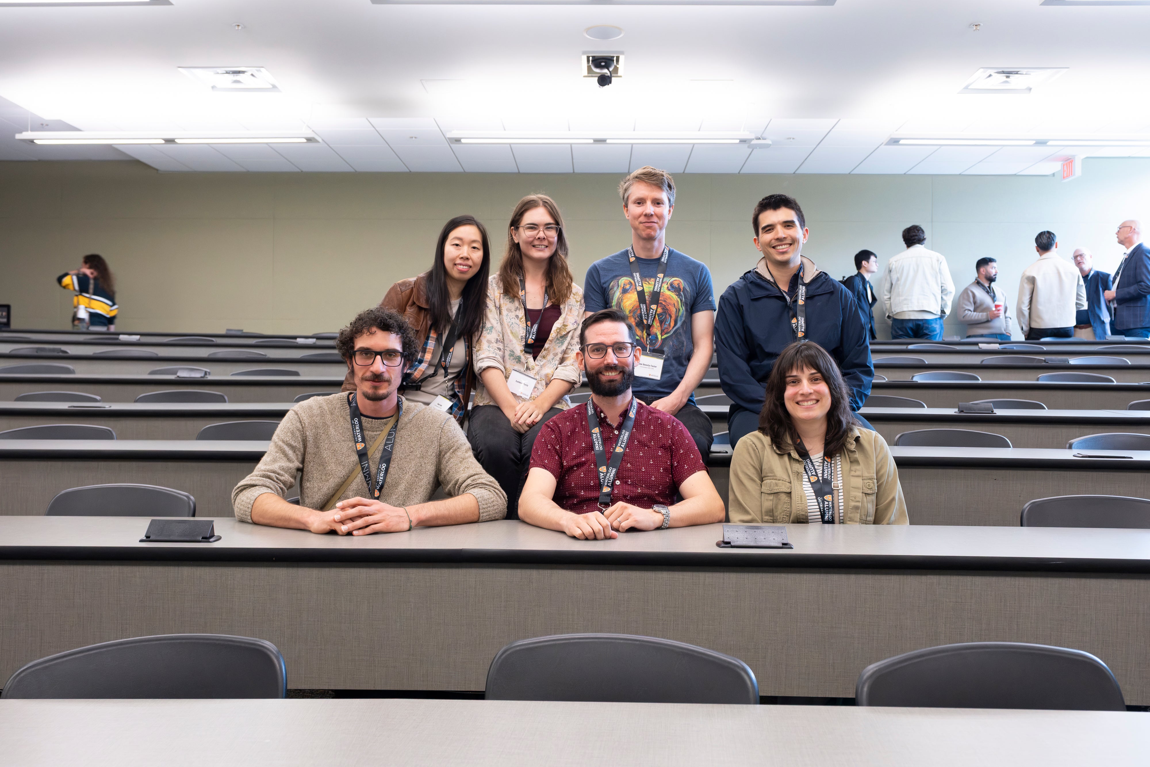 A group of seven people posing together in a lecture hall, with seats arranged in a grid behind them.