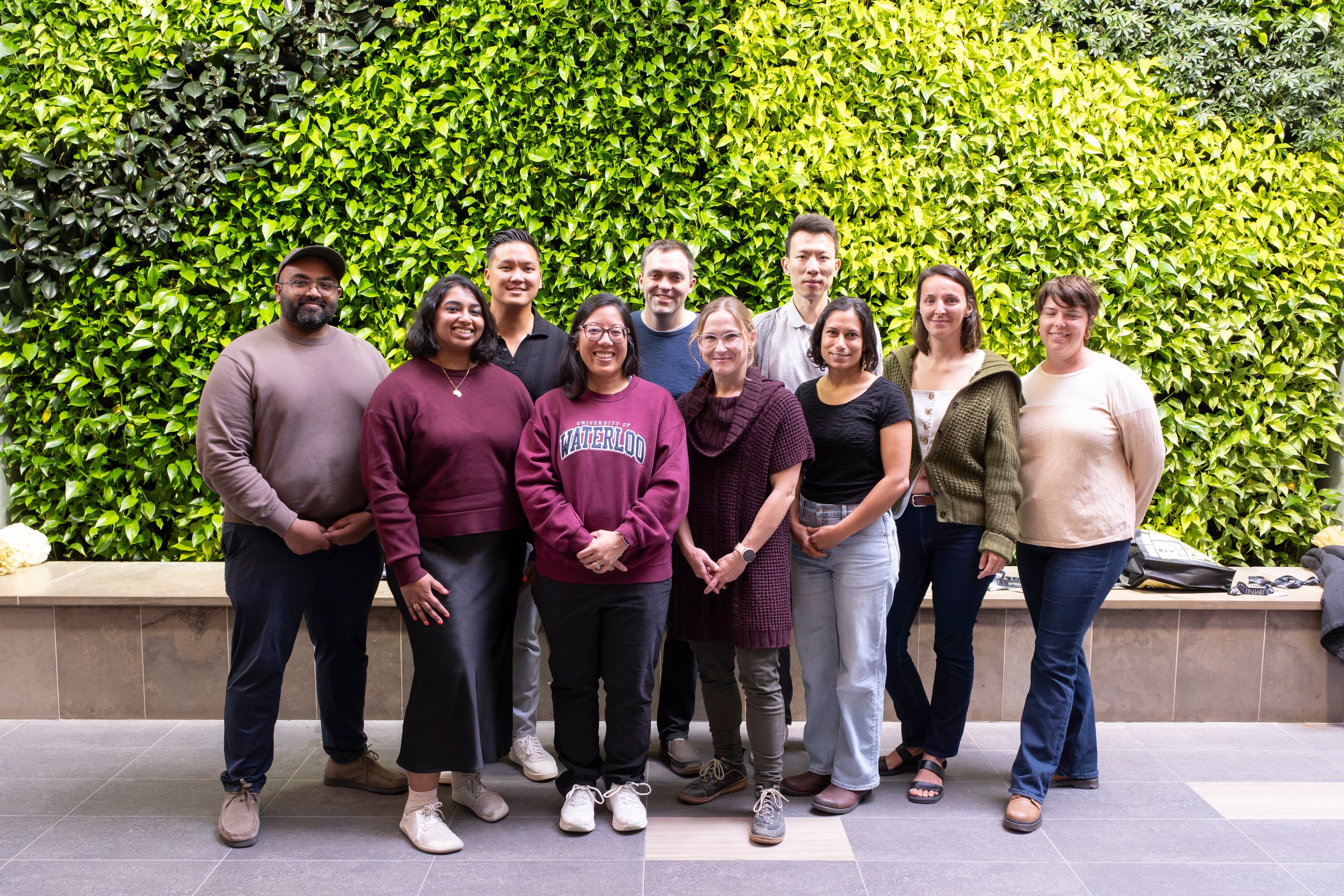 Ten people gather for a group photo in front of a green, living wall. 