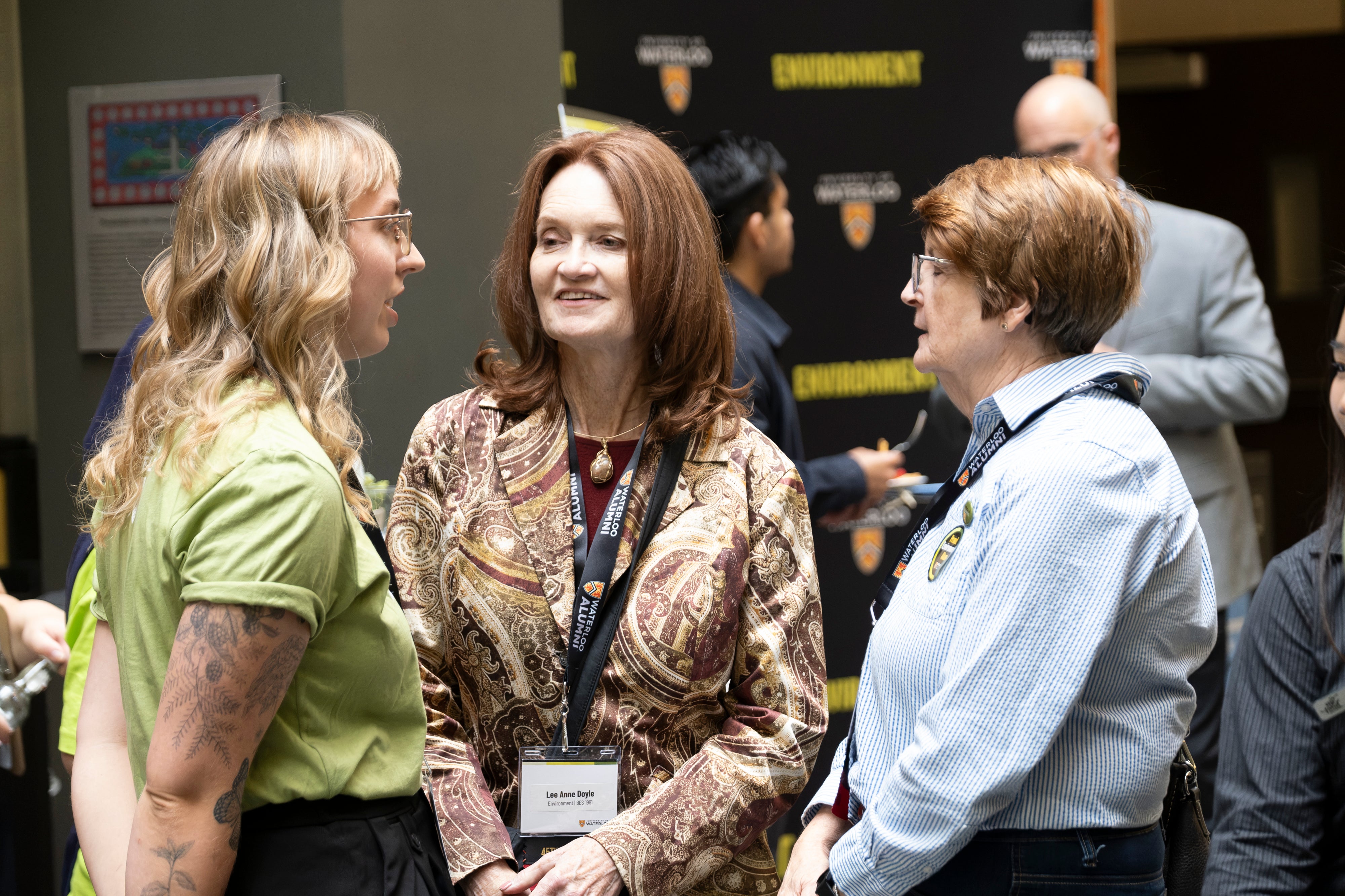 Three women engaged in conversation, with one wearing a green shirt and tattoos, the second in a patterned top, and the third in a blue striped shirt, all in a public setting.