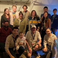 Thirteen people, including one holding a baby, pose indoors in casual clothing, smiling at the camera under soft lighting.