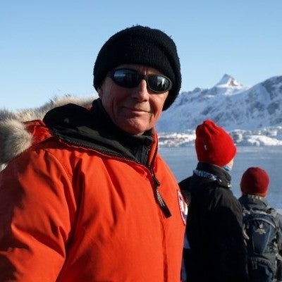 Man in red winter coat, black touque and sunglasses smiles with a wintery mountainous background.