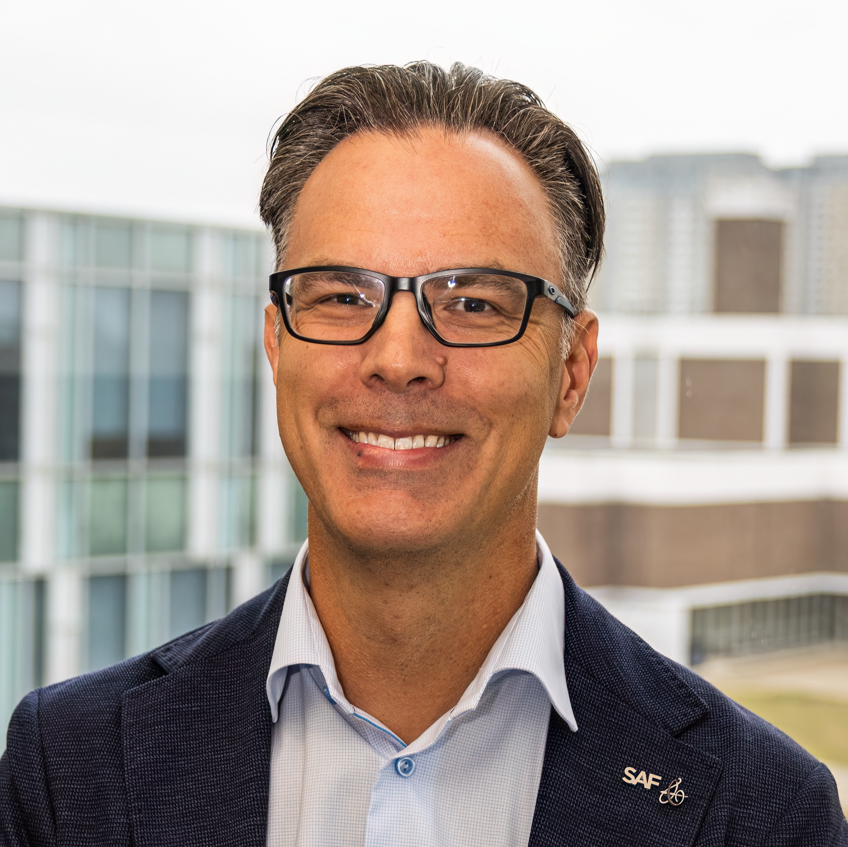 smiling man with black glasses in  baby blue collared shirt and dark blue blazer