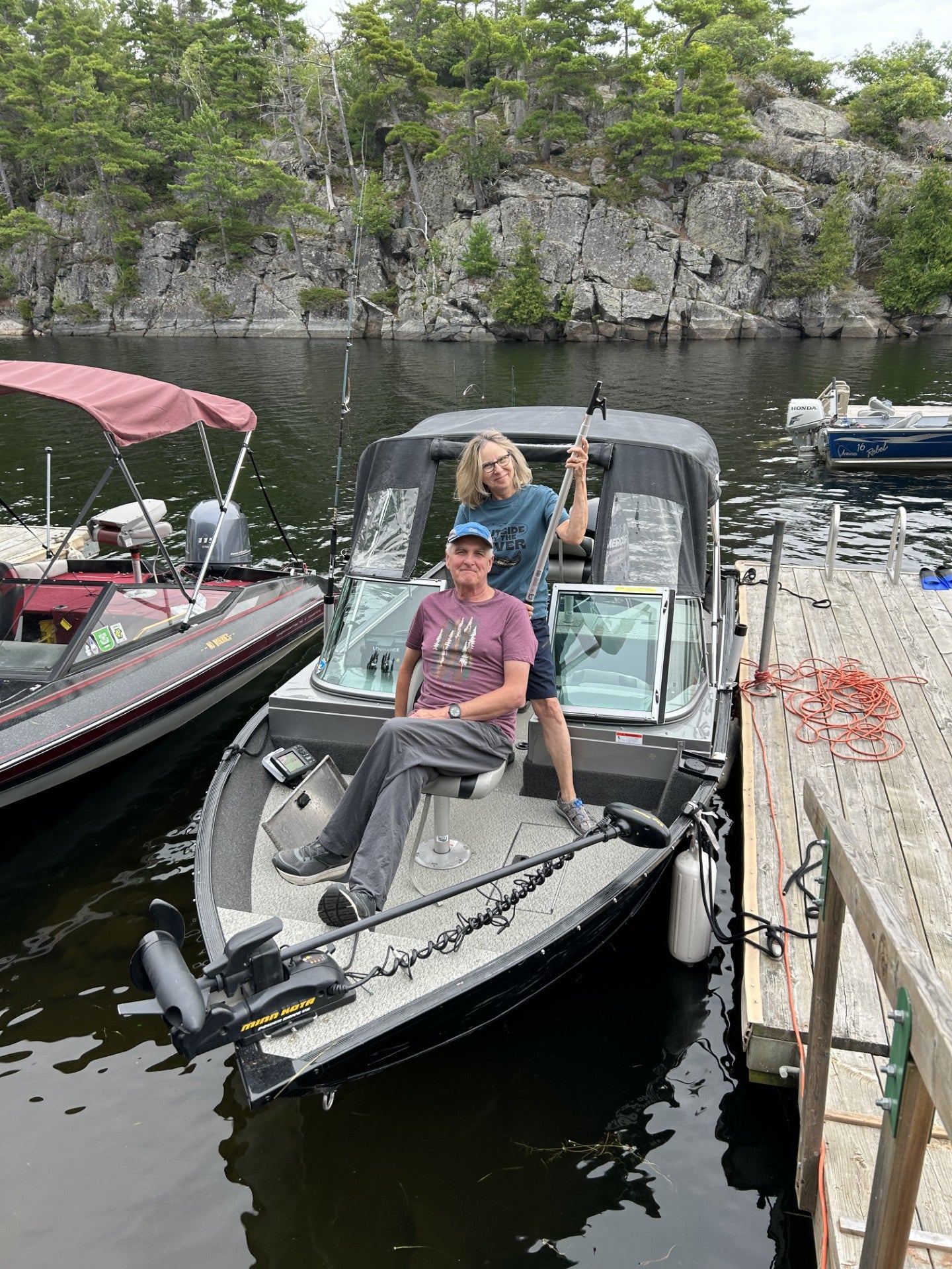 Man in ballcap and woman in blue tshirt smile from a fishing boat at a dock.