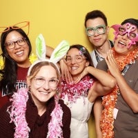 Five people wearing festive party accessories pose against a yellow backdrop, with colorful feather boas and oversized novelty glasses.
