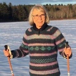 Woman with grey hair and glasses holds ski poles with snow in background.