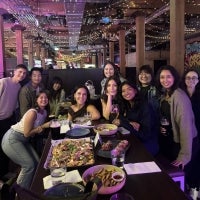 A large group of diverse individuals, appearing to be students or colleagues, pose for a candid group photo in a modern restaurant with high ceilings and string lighting.