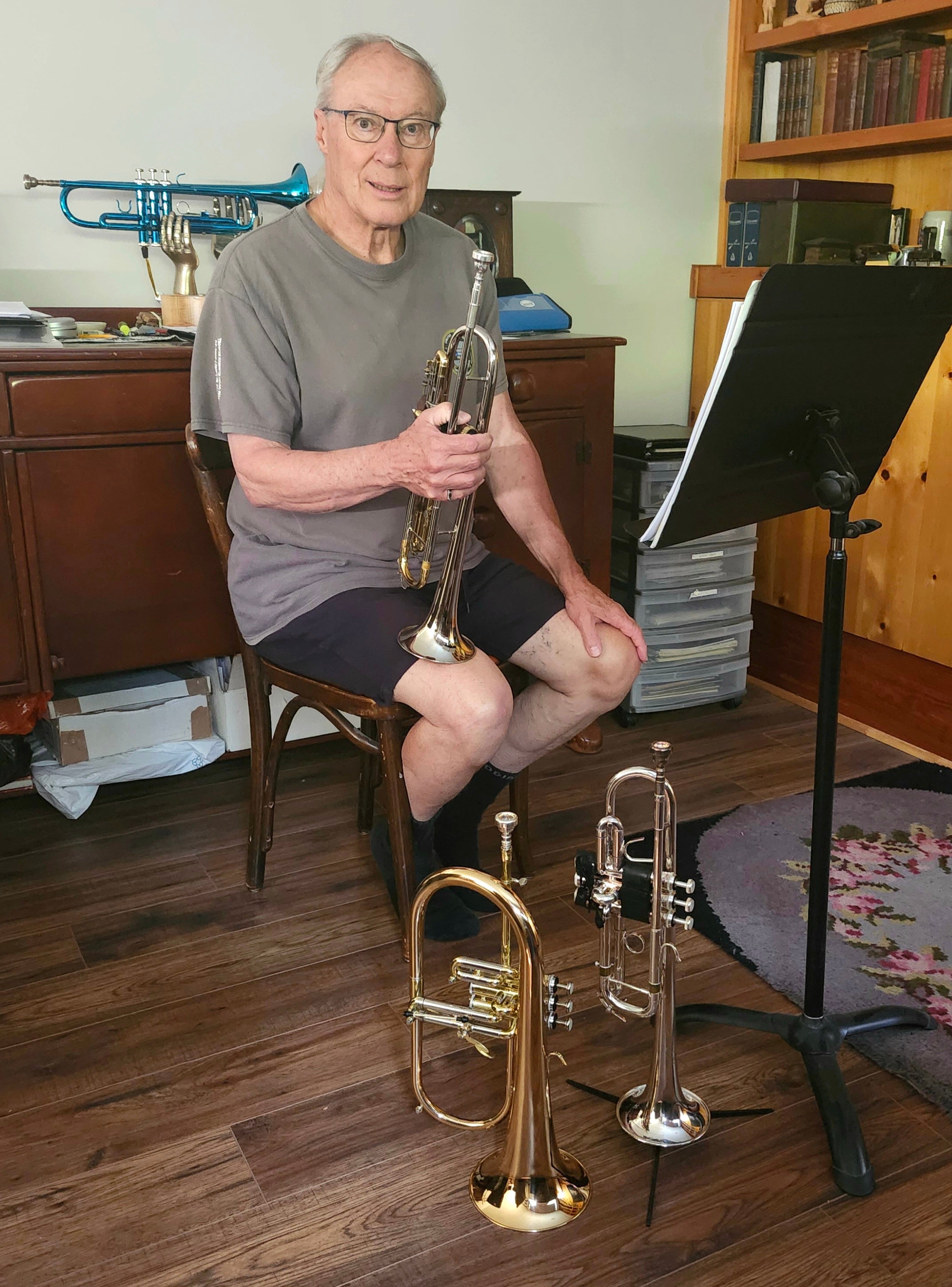 Man sits on stool with a trumpet in his hand and two trumpets on the ground. 