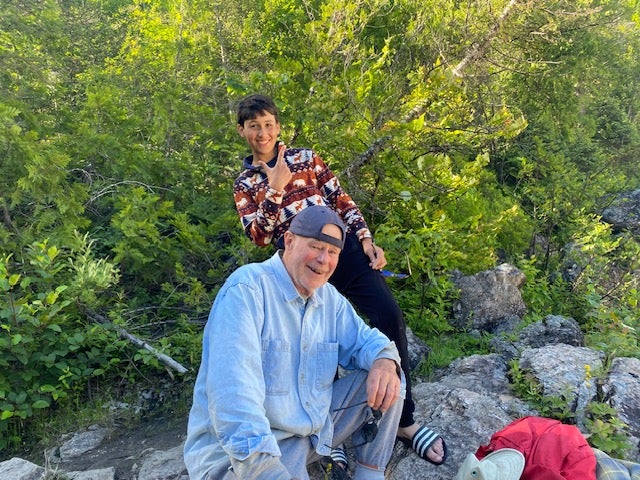 A smiling man in a light blue shirt and cap sits on rocks beside a younger person in a patterned shirt, surrounded by green trees.