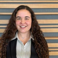 A woman with long curly hair wearing a black blazer over a striped shirt stands in front of a wood slat wall.