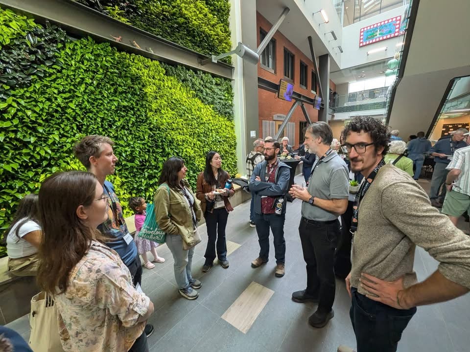 A group of seven people talking at a reception with high ceiling and a green plant wall. 