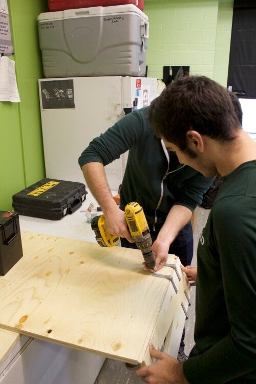 One student drilling hinge bracket to attach two pieces of wood, while another student holds on of the pieces