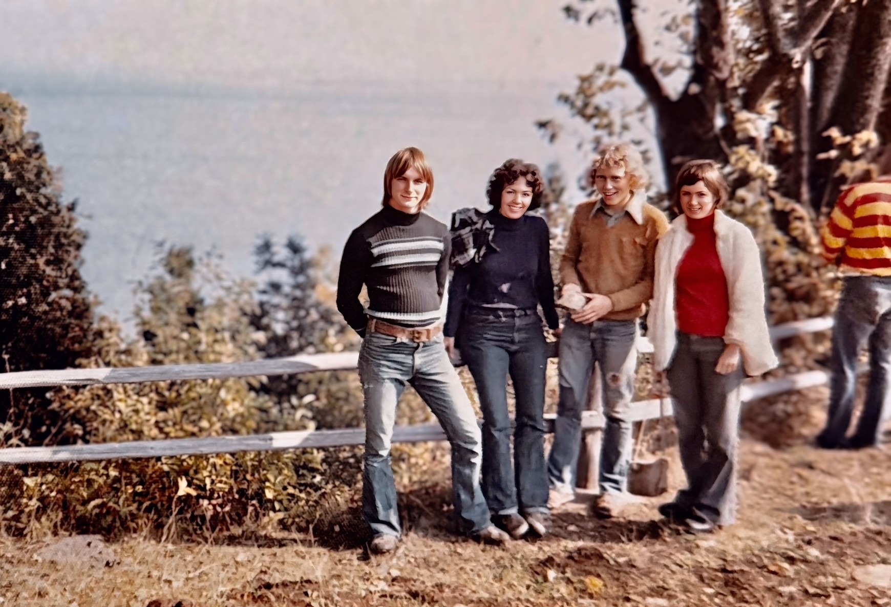 Four young people standing in a row outdoors by a wooden fence near a lake, dressed in 1970s-style sweaters, blue jeans, and a white fur jacket.