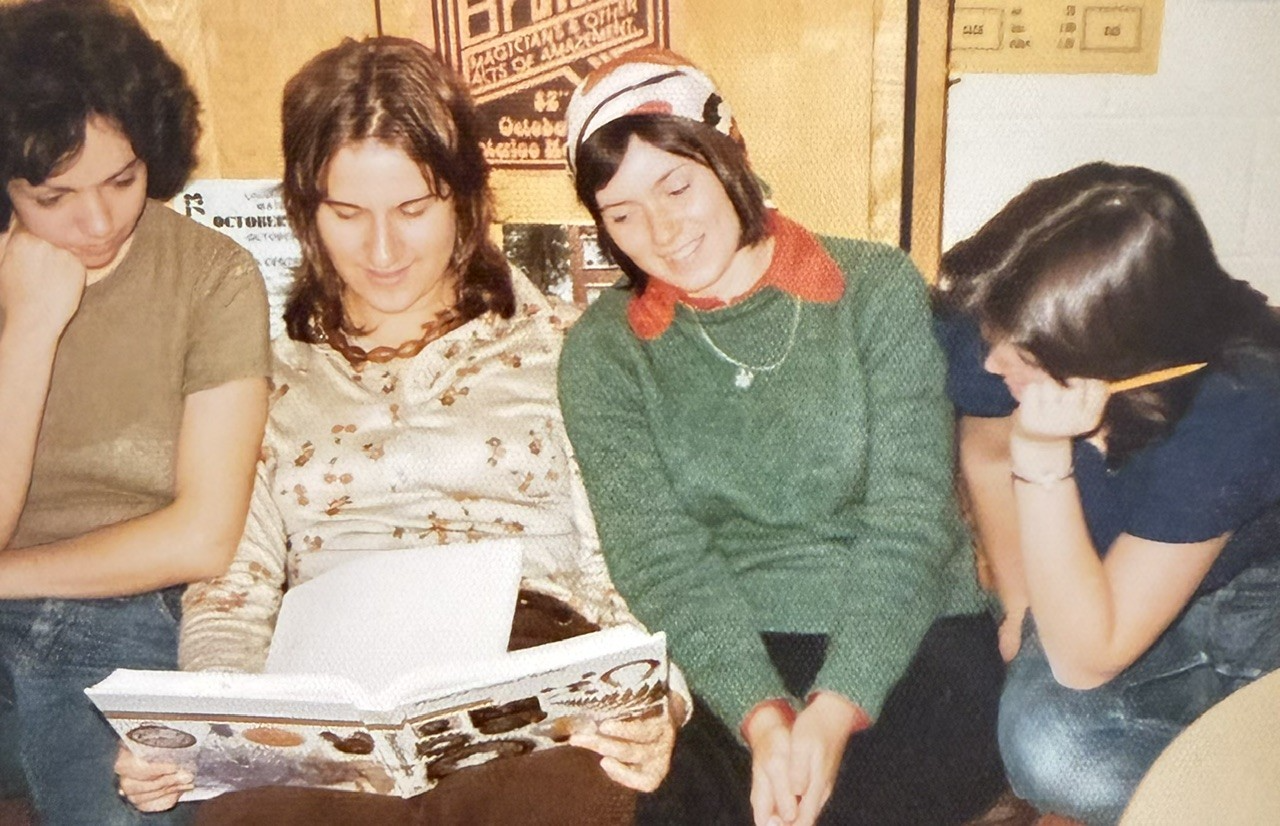 Four young women sit together, with two looking at an open book, one wearing a headscarf and green sweater, and another holding a yellow pencil.