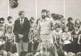 Black and white photo of man in striped shirt speaking in a group of children. 
