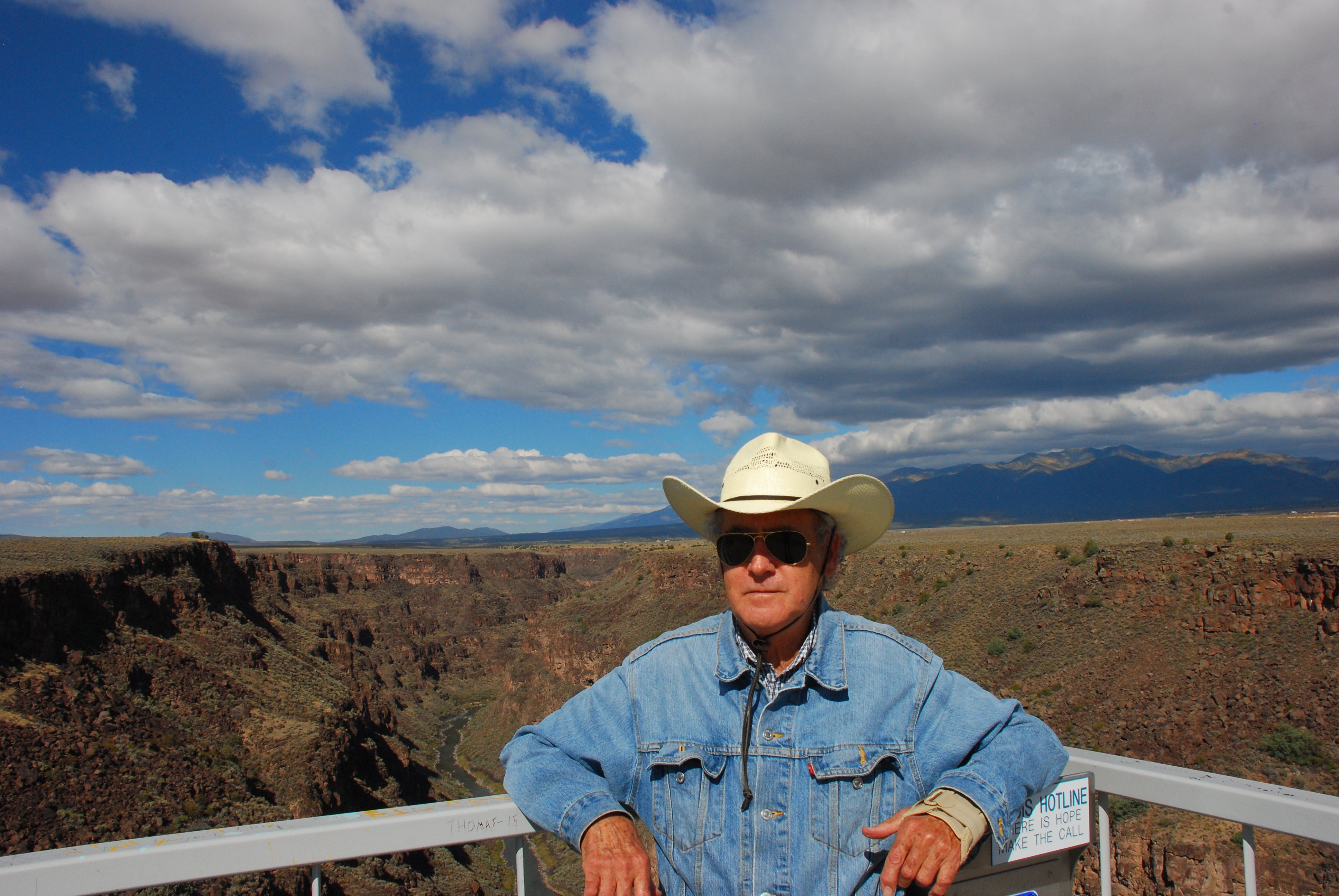 Man in denim shirt and white cowboy had leans against railing with prairies in the background. 