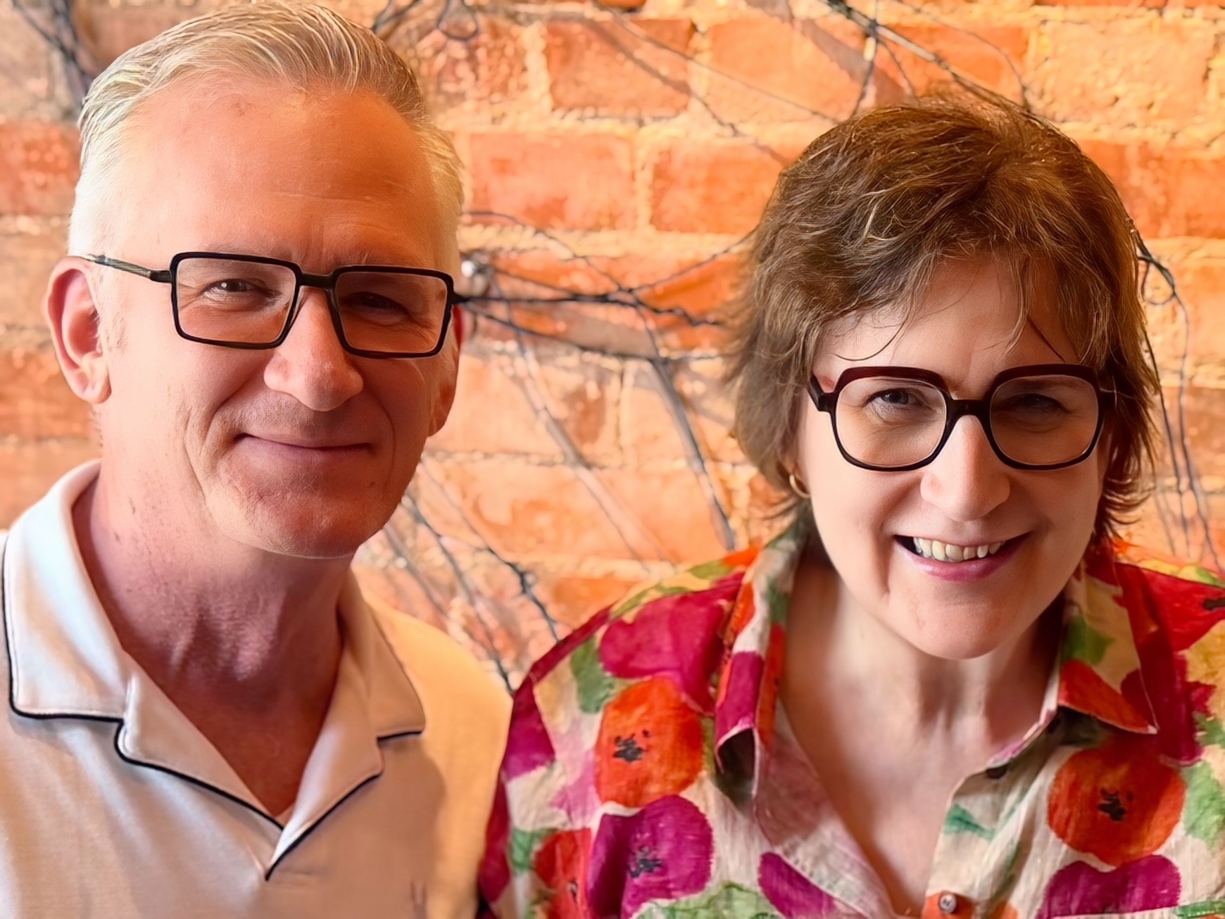 Man and woman, both in glasses smile in front of a brick wall.