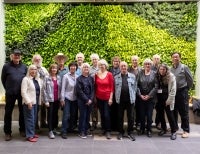 A diverse group of individuals smile, lined up in front of a large plant wall.