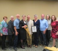 A group of eight adults stand together indoors against a beige wall, with one man holding a framed plaque.
