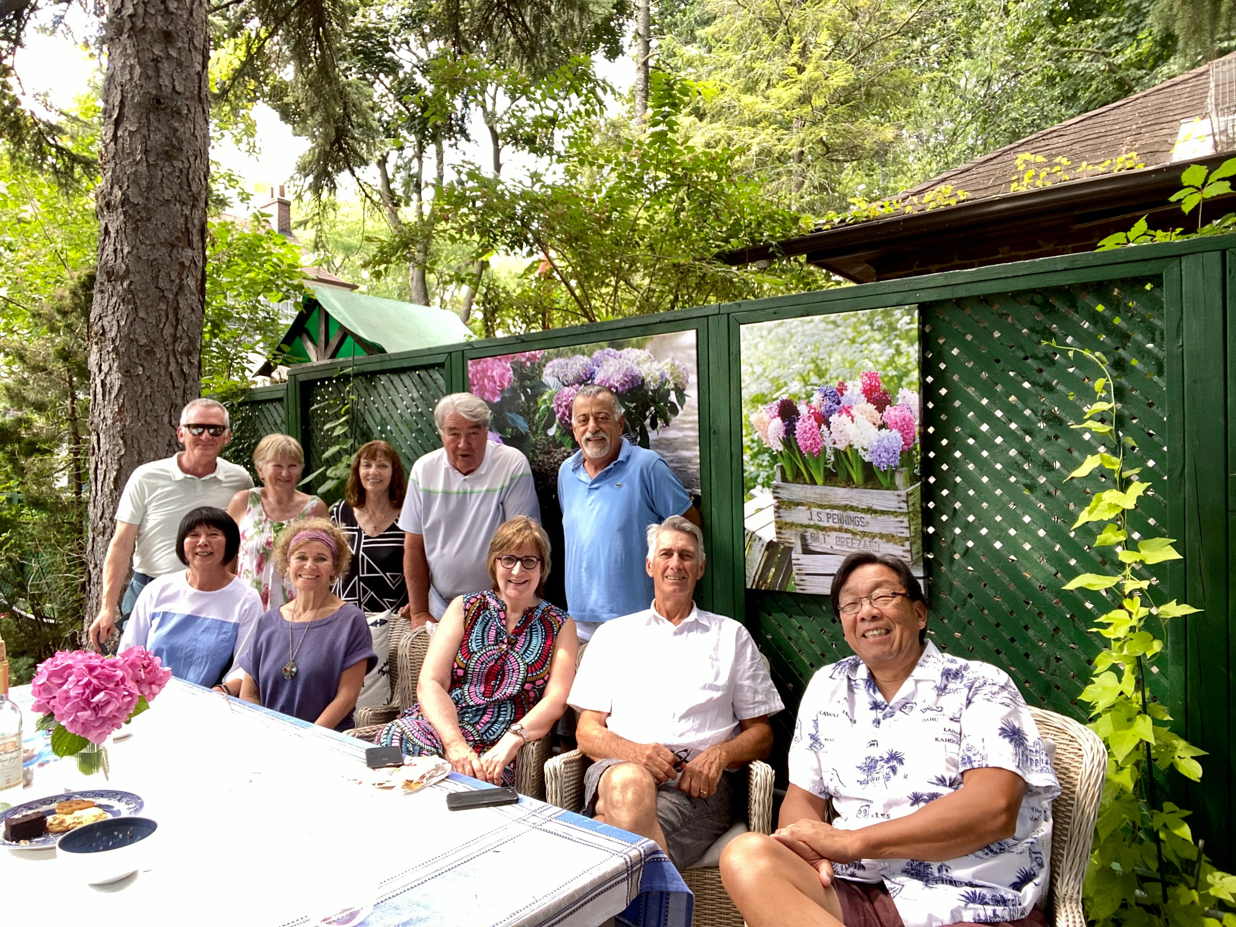Group of people at a back yard table sit at table, with others standing behind them. 
