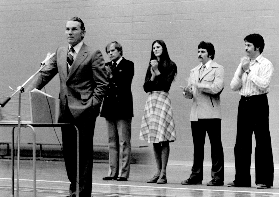 Man in suit speaks into microphone at podium while four students behind clap (black and white, 1977)
