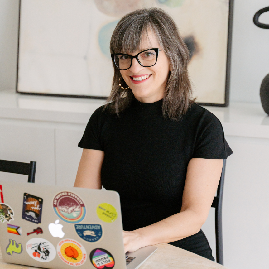 A woman with black glasses and gray hair sits at a table with a sticker-covered laptop, in front of framed wall art.