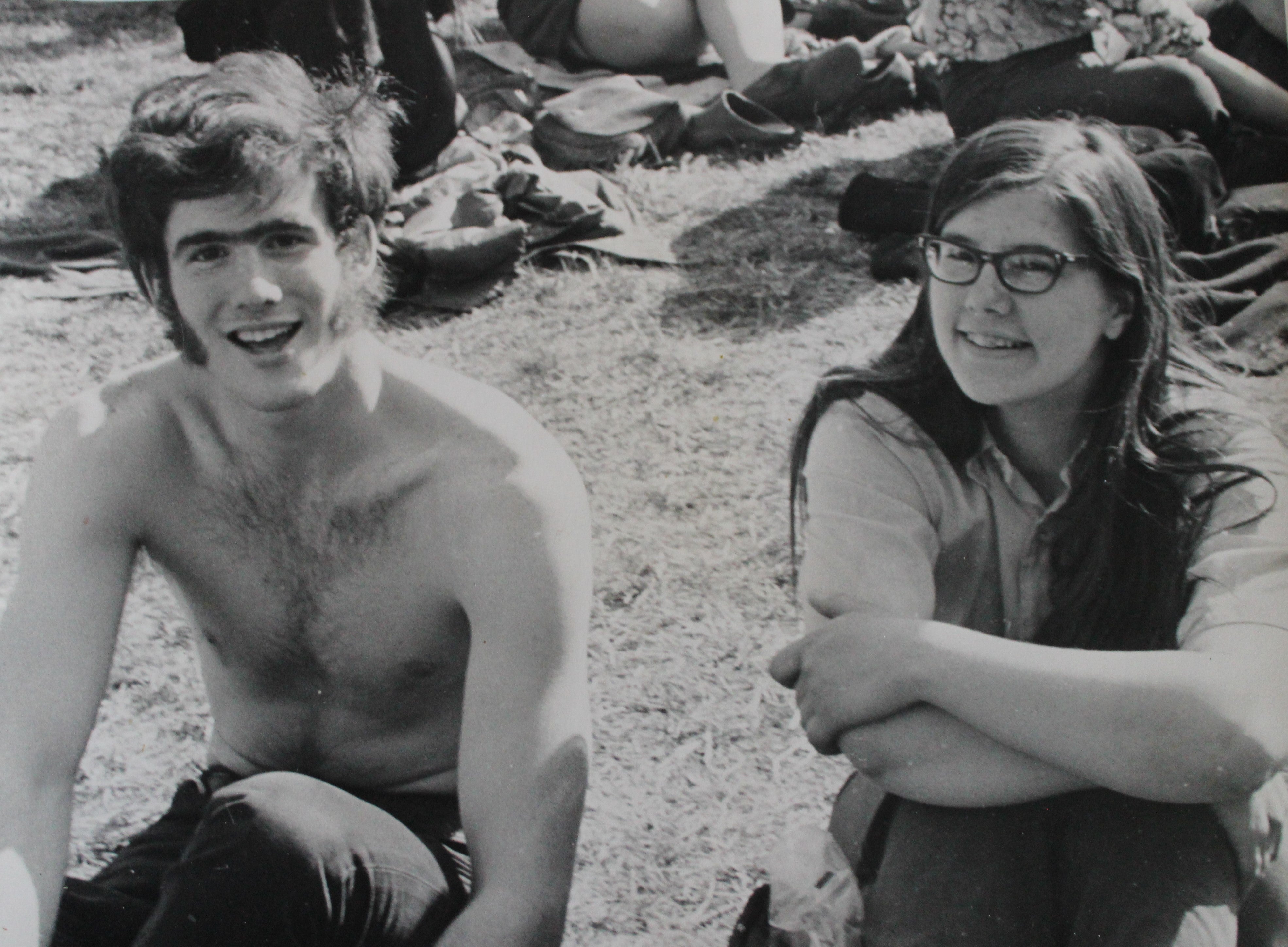 A black and white photo of a shirtless young man with sideburns and a young woman with long hair and glasses sitting together on the grassy ground.