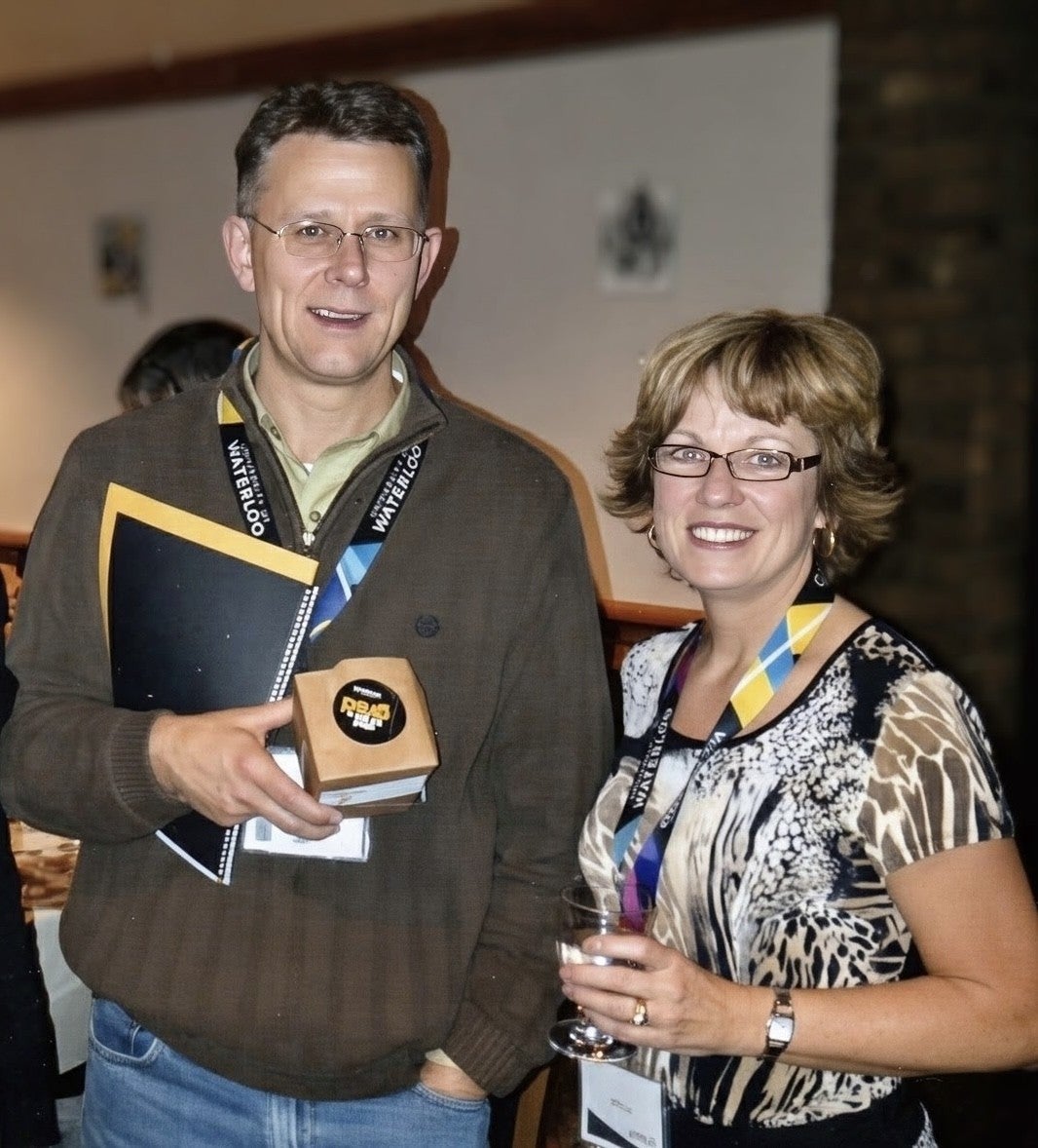 A man holding a notebook and a small box and a woman holding a glass are smiling at the camera.
