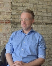 Man in glasses wearing a blue button-up shirt sits against a brick wall, posing for a portrait.