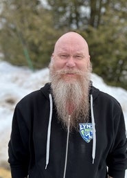 Bald man with a long gray beard, wearing a black zip hoodie with a blue Lynx patch, outdoors in a snowy wooded setting.