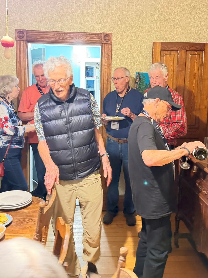Several older adults gather indoors, with one man in a puffer vest in the foreground, and another man in a black cap pouring wine into a glass.