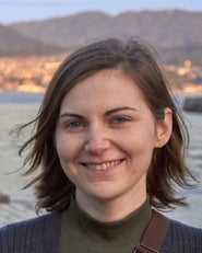 A smiling woman with short brown hair is shown in a close-up outdoors, with water and mountains in the background.