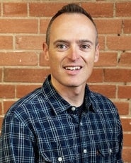 A man with short brown hair and a plaid shirt smiles against a brick wall