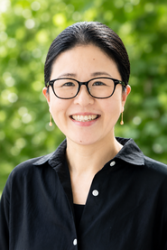 Smiling woman with black hair and glasses, wearing a black button-down shirt and gold drop earrings, outdoors.