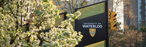 	a photo of the University of Waterloo sign at the bottom of ring road partially obscured by a tree.