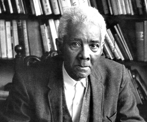 A black and white photo of CLR James, an older black man wearing a suit jacket in front of a shelf of books.